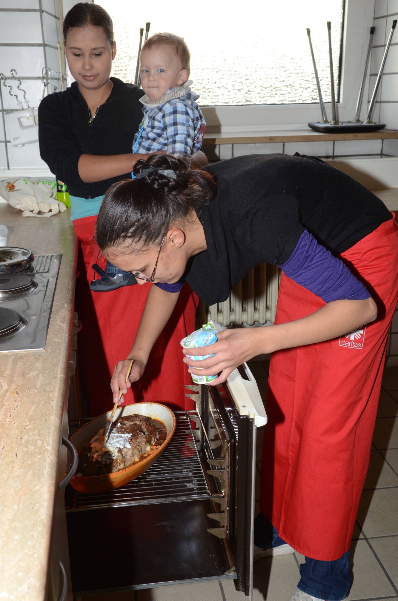 Eine junge Frau bestreicht am Backofen einen Braten, eine andere sieht mit Kind auf dem Arm zu.